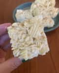 Close-up of a hand holding a square rice cereal treat with a plate of treats in the background.