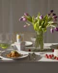 Elegant table setting with yellow and purple tulips, pasta dishes, baguette, and drinks on a white tablecloth.
