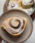 Cinnamon roll with icing on a beige plate, with more rolls in a pan in the background.