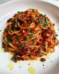 Spaghetti with tomato sauce, capers, olives, and fresh parsley on a white plate.