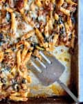Baked penne pasta with melted cheese, spinach, and sausage on a baking sheet with a metal spatula.