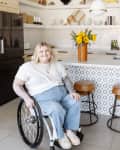 Person in a wheelchair in a modern kitchen with open shelves, sunflowers on the counter, and bar stools.