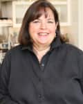 Woman in a black shirt smiling in a kitchen with bowls of fruit and utensils.