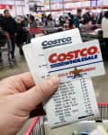 Hand holding Costco membership card and receipt in a busy store checkout line.
