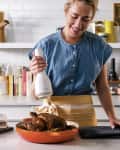 Woman in a kitchen seasoning a roasted chicken in an orange pan, surrounded by spices and kitchen decor.
