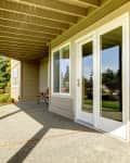 Covered patio with beige siding, white French doors, and a view of a green lawn and trees.