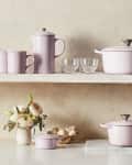 Pastel pink cookware set on shelves with mugs, cutting board, and flowers.
