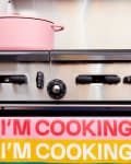 Pink pot on stove with colorful towel reading "I'm Cooking" in bold letters.