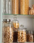 Pantry shelf with glass jars of pasta, cereals, and snacks, alongside bottled water and a paper bag.