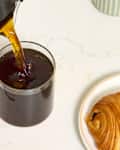 angled shot of someone pouring coffee into a glass mug, with a chocolate croissant next to the coffee