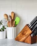 Wooden knife block with black-handled knives on a kitchen counter, surrounded by potted plants and utensils.
