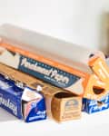 Parchment paper and aluminum foil boxes on a kitchen shelf next to a glass jar and bottles.