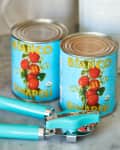 Two cans of Bianco DiNapoli crushed tomatoes on a marble counter with a turquoise can opener.