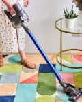 Person vacuuming a colorful geometric rug next to a blue chair and a round table with plants.