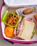 Ham sandwich, pretzels, orange, almond butter, apple sauce, and orange juice in a pink lunchbox.
