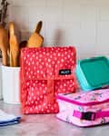 Colorful lunch bags and containers on a kitchen counter with wooden utensils and potted plants in the background.