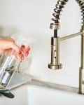Hand holding a spray bottle near a stainless steel kitchen faucet.
