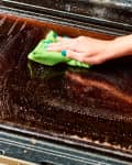Hand cleaning an oven door with a green cloth and soapy water.