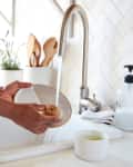 Person washing a plate under a kitchen faucet with soap dispenser and wooden utensils nearby.