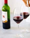 Two glasses of red wine on a marble countertop with a wine bottle and potted plant in the background.