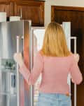 Woman in pink sweater opening stainless steel refrigerator in kitchen with wooden cabinets and potted plant.