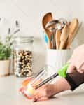 Hand lighting a candle in a glass jar on a kitchen counter with utensils and a plant in the background.