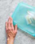 Hands cleaning a blue silicone food storage bag with a cloth on a marble surface.