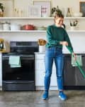 A woman standing in a white kitchen, sweeping the concrete floors with a broom