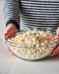 Person holding a glass bowl filled with freshly popped popcorn on a marble countertop.