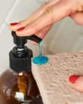 Hand dispensing blue liquid soap onto a sponge from a pump bottle.