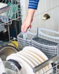 Person loading a dishwasher with plates, bowls, and a cutlery basket, wearing a blue striped shirt and jeans.