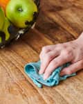 Hand cleaning a wooden table with a blue cloth and spray bottle, next to a fruit bowl with green apples.