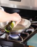 Pouring olive oil into a stainless steel skillet, a bowl of white vinegar and bottle of vinegar next to the stove