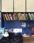 Kitchen with blue cabinets, books on a shelf, knives on a magnetic strip, fruit basket, and colorful wall decor.