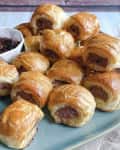 a platter of small sausage rolls on a square turquoise plate with a brownish-red dipping sauce