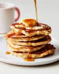 Stack of five oatmeal pancakes in a white plate, topped with a pat of butter, maple syrup being pored on top