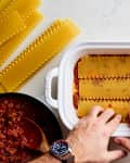 Hands assembling lasagna in a white dish with pasta sheets, meat sauce, ricotta, and shredded cheese on a marble surface.