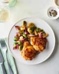 Roasted chicken thighs with Brussels sprouts, apples, and red onions on a white plate, next to a glass of white wine.