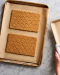 Gingerbread house pieces on baking sheets, including roof with scallop pattern and walls with window and door outlines.