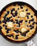 Pancake is baked and served with butter, maple syrup, and extra blueberries.