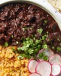 Overhead view of chile colorado and rice in a green bowl topped with radishes and cilantro. Half a lime in the top left corner of the shot, and 3 tortillas peeking in on the top right corner of the shot.