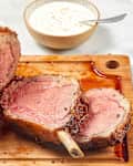 Angled view of prime rib resting on a wooden cutting board, first two slices are cut and theres a bowl of horseradish cream sauce behind it.