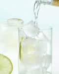 Front view of two gin and tonics in high ball glasses, each with a lime in them and tonic being poured into the front glass.