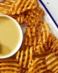 Overhead shot of waffle fries, and a small white bowl of chick fil a sauce on a white dish with a blue rim. In the bottom left of the image theres a hand dipping a waffle fry into the sauce.