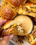close up shot of a tray of cheeseburger sliders in a white pan, with one slider being dipped into the side of sauce on the pan.