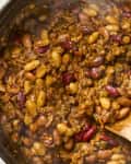 overhead shot of cowboy beans in a large dutch oven with a wooden spoon stirring the beans.