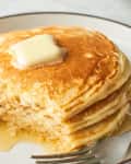 Fluffy pancakes stacked on a plate, topped with a pat of butter and drizzled with syrup.