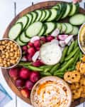 Cucumber slices, radishes, snap peas, hummus, and pretzel crisps on a wooden board with dips.