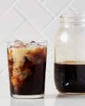 Iced coffee with cream swirling in a glass next to a mason jar of cold brew on a white tile background.