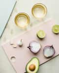 Hands slicing lime on a blue cutting board with avocado, garlic, onion, and lime on pink board, next to two glasses.